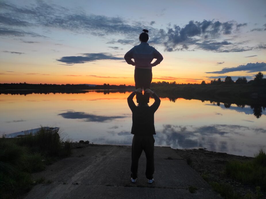 Duo Circ2M:n taiteilijat seisovat järven rannalla toinen toisen olkapäillä selät kohti kameraa. He katsovat horisontissa näkyvää auringonlaskua. Pilvet heijastuvat tyynen veden pintaan.