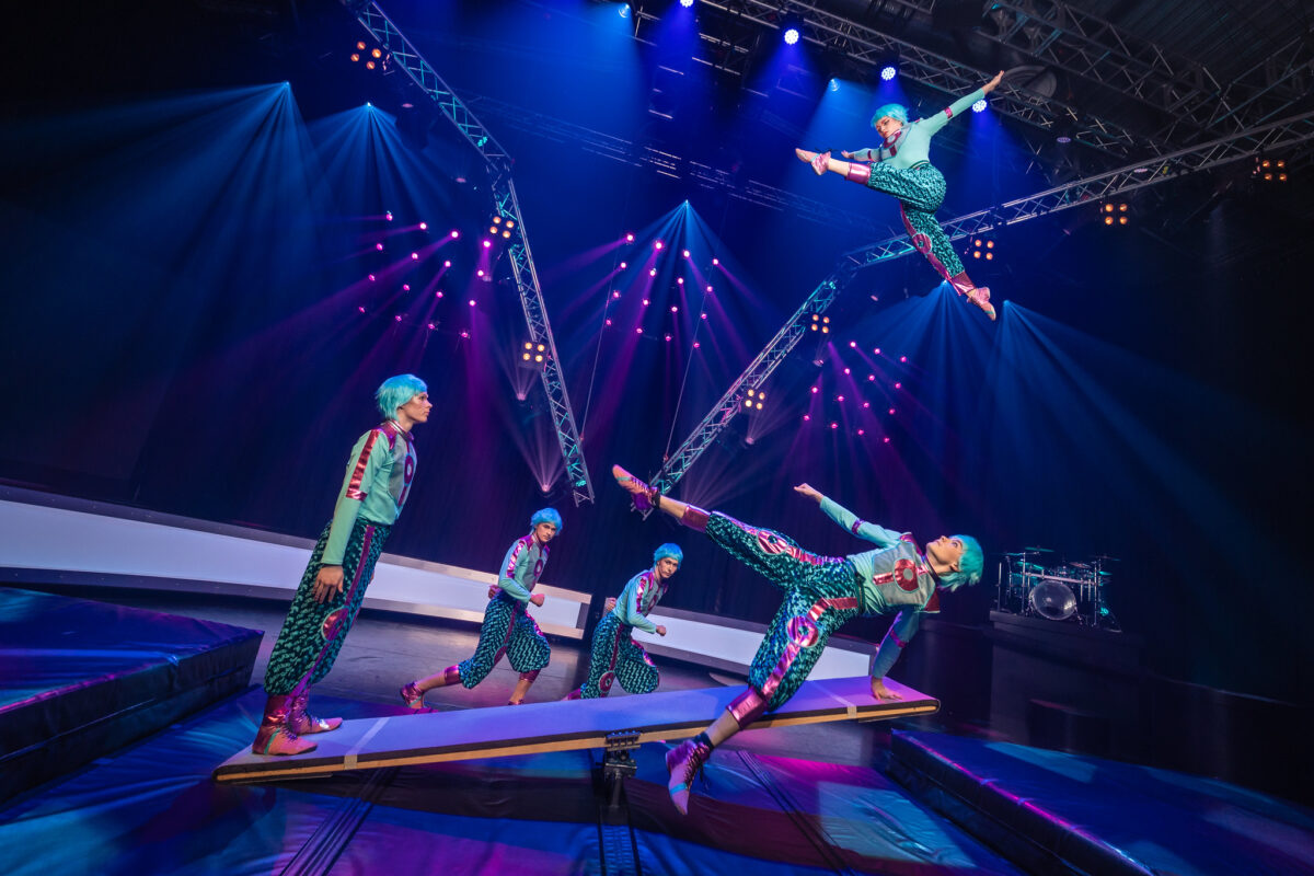 Viisi turkooseihin asuihin pukeutunutta sirkusartistia taituroi vipulaudalla.